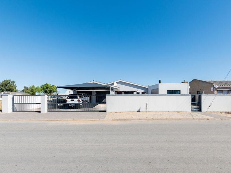 Front view of the property featuring a secure perimeter wall, sliding gate, pedestrian entrance, and a covered carport area.