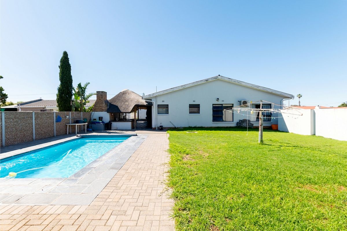 Rear exterior view featuring a swimming pool, paved patio area, thatched lapa, and a spacious lawn enclosed by boundary walls.