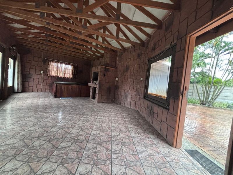 Spacious covered patio featuring exposed timber roof trusses, stone-clad walls, tiled flooring, and a built-in braai, offering direct access to the garden.