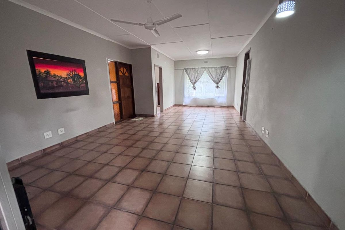 A spacious, empty living area featuring neutral walls, tiled flooring, a ceiling fan, and natural light from a large window.