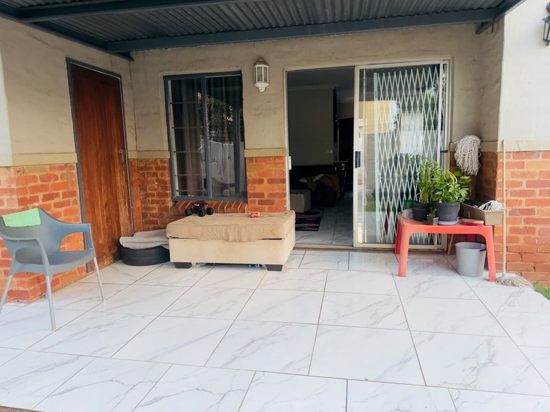 A covered patio area featuring marble-look tiled flooring, brick accents, and a sliding glass door with a security gate providing access to the interior.