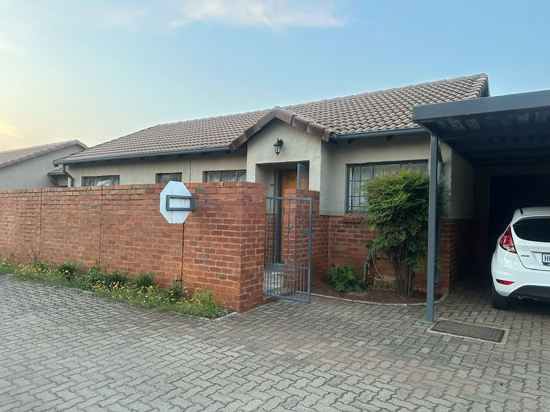 Front elevation of a townhouse featuring a brick boundary wall, tiled roof, and a covered carport with paved driveway access.
