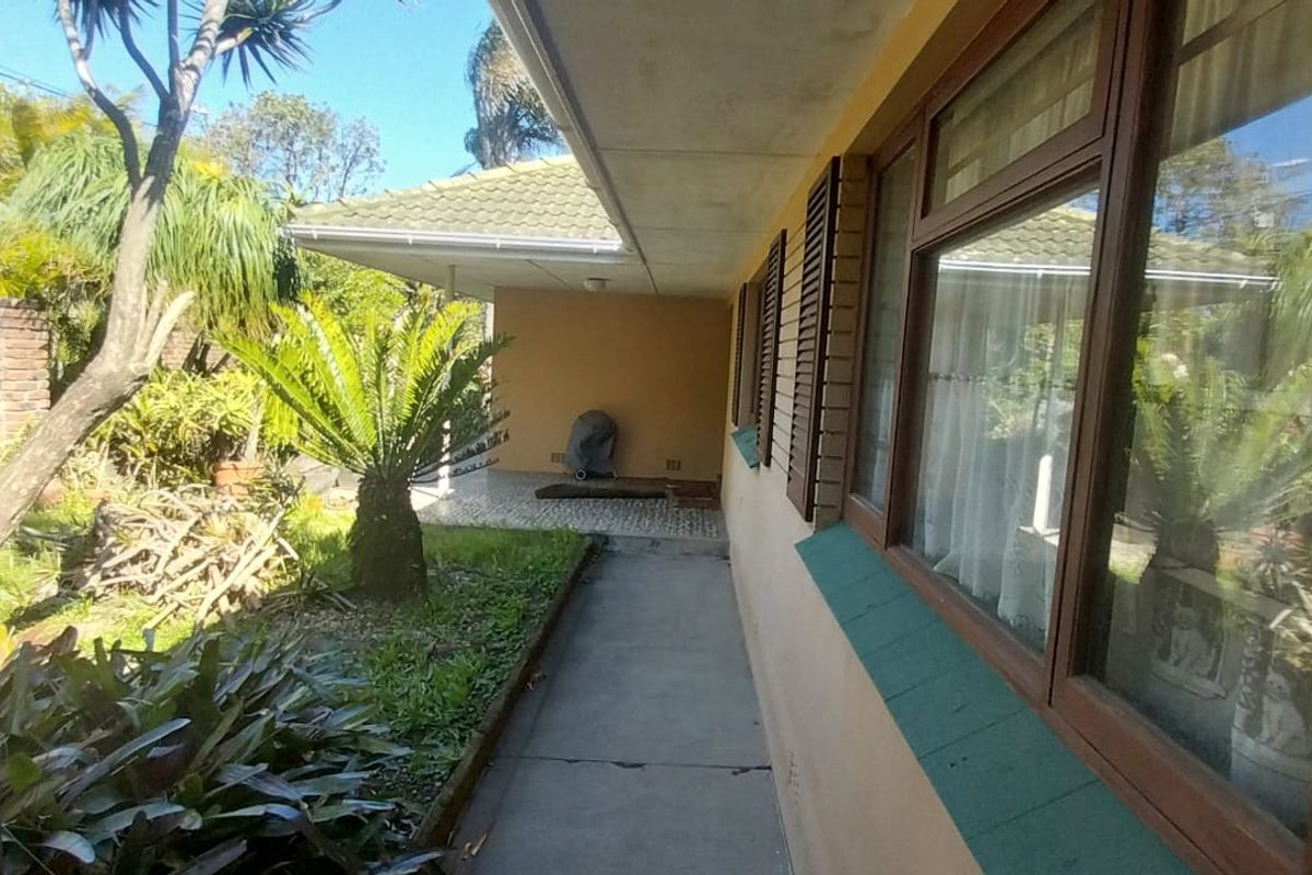 A rear exterior view featuring a covered patio, concrete walkway, and established garden with brick boundary walls and wooden window shutters.