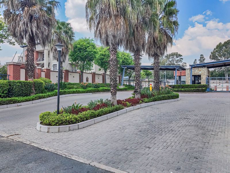 A well-maintained apartment complex entrance featuring a paved driveway, landscaped garden beds, and secure gated access with covered parking structures.
