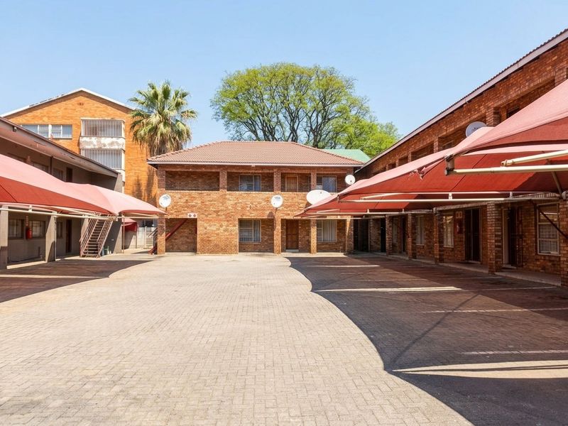 Exterior view of an apartment complex featuring a paved courtyard, brick building facades, and covered parking bays with shade cloth structures.