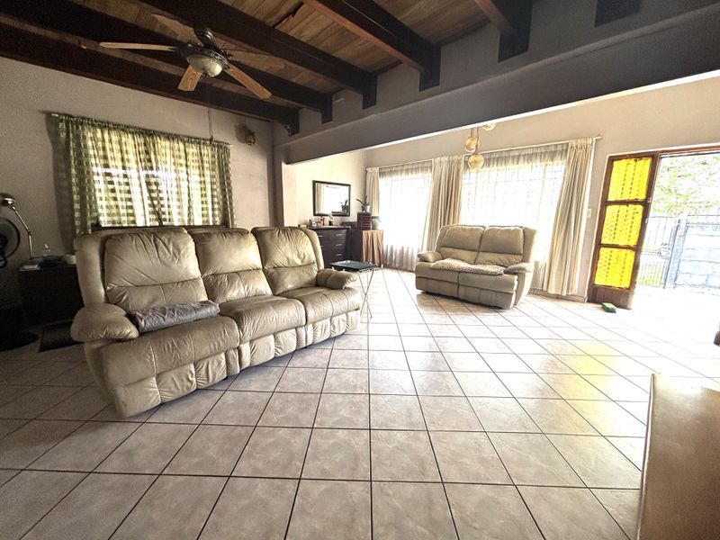 Spacious living room featuring exposed timber ceiling beams, tiled flooring, and natural light through large windows with access to an exterior door.