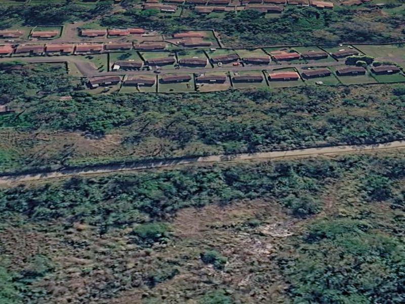 An aerial view of a vacant residential plot featuring natural vegetation and access to nearby established housing developments.