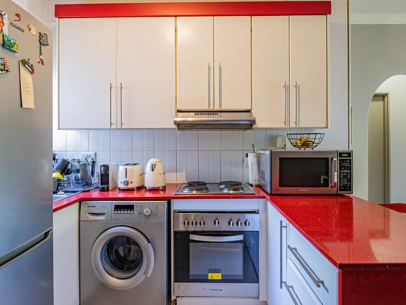A functional kitchen featuring white cabinetry, a vibrant red stone countertop, tiled backsplash, and built-in appliances including an oven, hob, and washing machine.