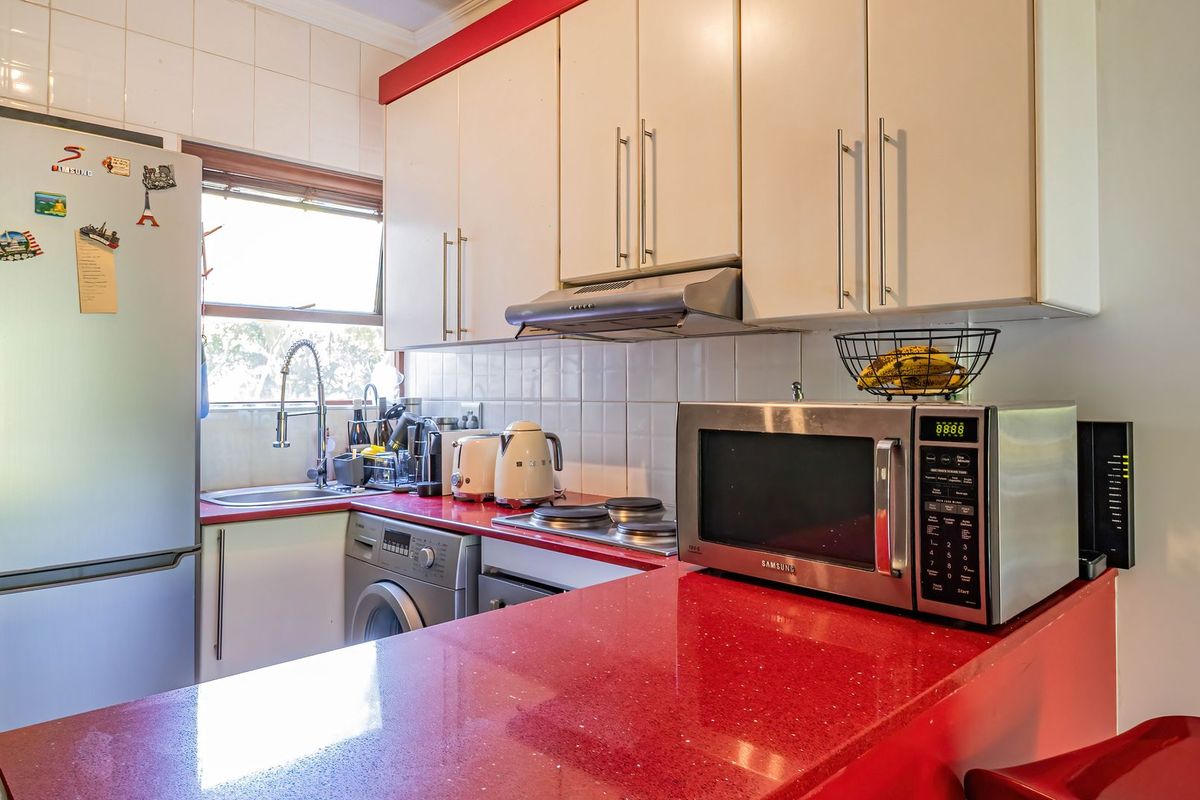 A compact kitchen featuring red stone countertops, white cabinetry, tiled backsplash, and integrated appliances including a washing machine and extractor fan.