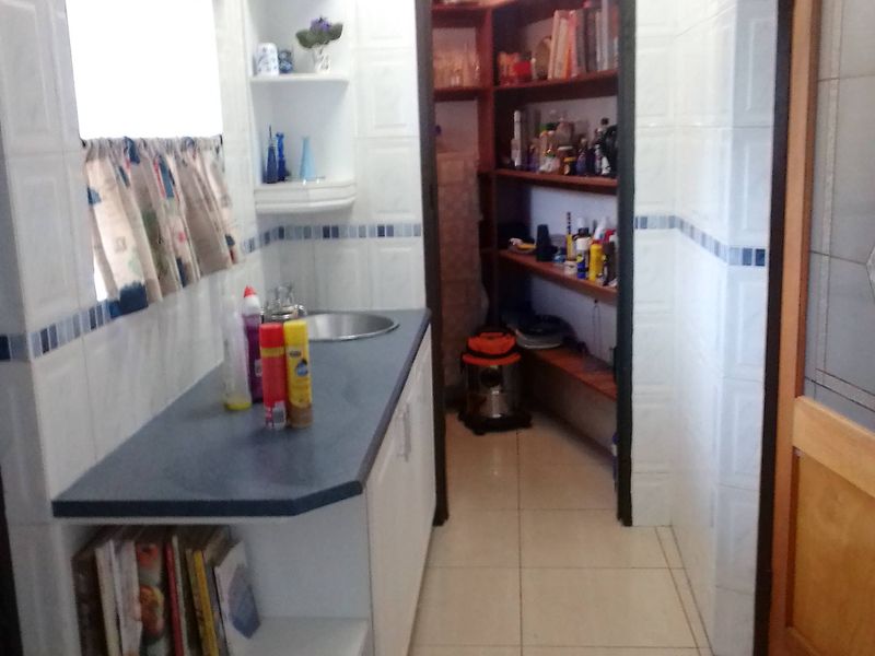 A functional scullery featuring tiled walls, built-in cabinetry with a laminate countertop, a stainless steel sink, and an adjoining walk-in pantry with shelving.
