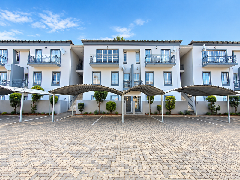 Front elevation of a modern apartment complex featuring paved parking areas, covered carports, and balconies with wrought iron railings.