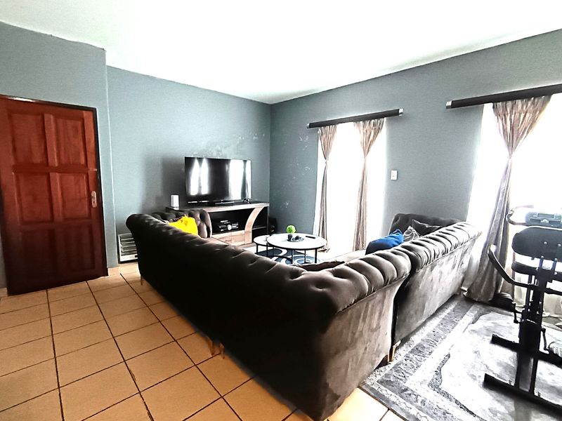 A living room featuring tiled flooring, a timber door, and large windows with curtain rails, currently furnished as a lounge area.