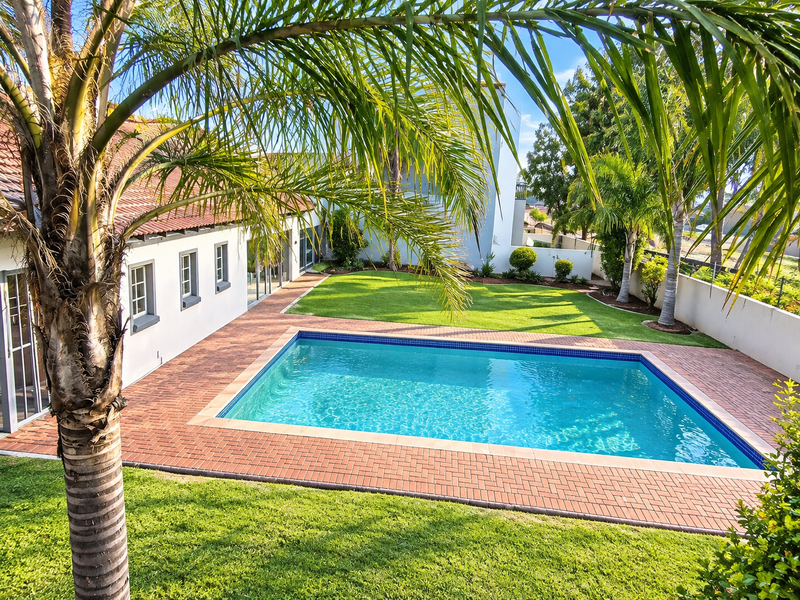 A well-maintained outdoor pool area featuring a clear swimming pool, brick paving, and a manicured lawn, set against the rear elevation of the property.