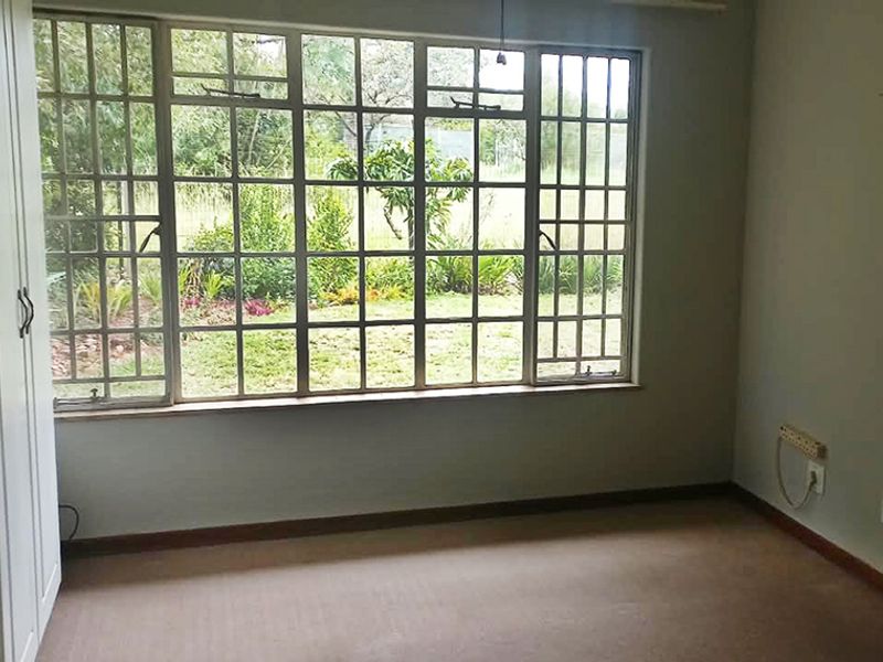 A bright, empty bedroom featuring built-in cupboards, carpeted flooring, and a large multi-pane window overlooking the garden.