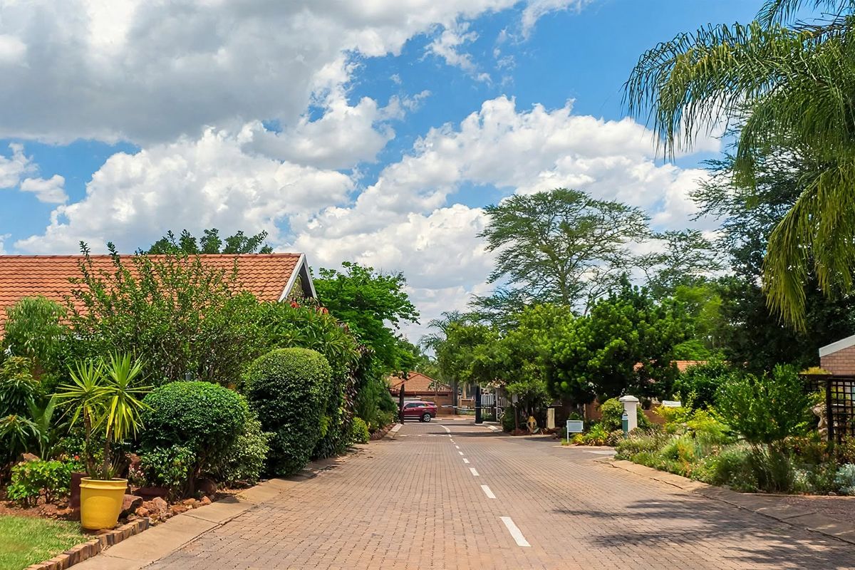A well-maintained paved street within a retirement village, featuring established landscaped gardens and brick residential buildings under a clear blue sky.