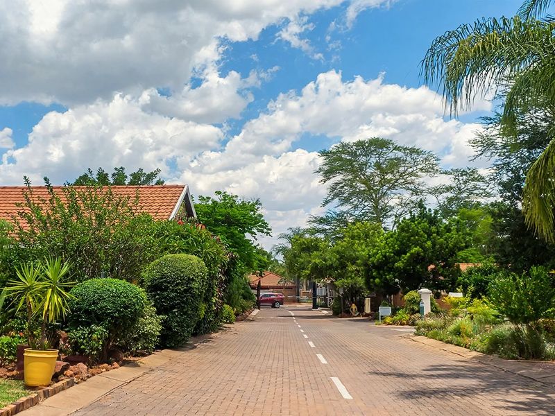 A well-maintained paved street within a retirement village, featuring established landscaped gardens and brick residential buildings under a clear blue sky.