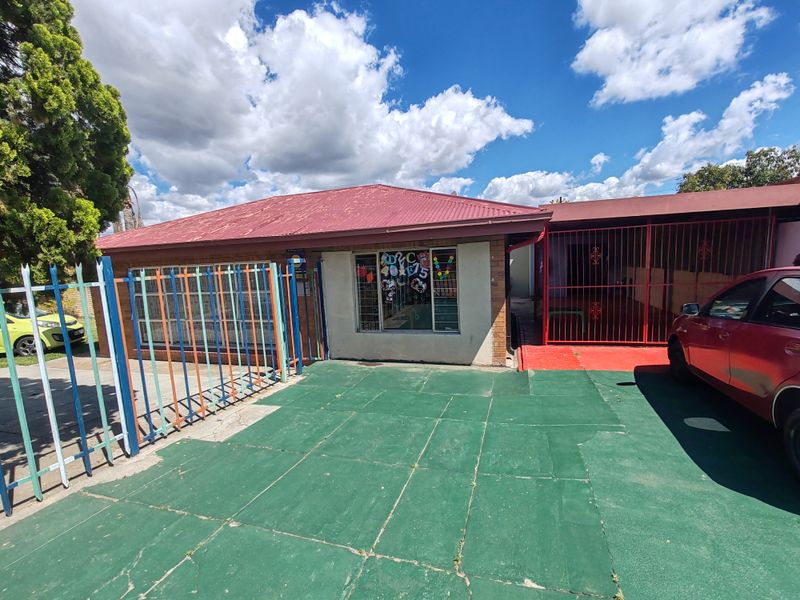 Front exterior view of a single-story house featuring a corrugated metal roof, brick facade, security gates, and a paved driveway area.