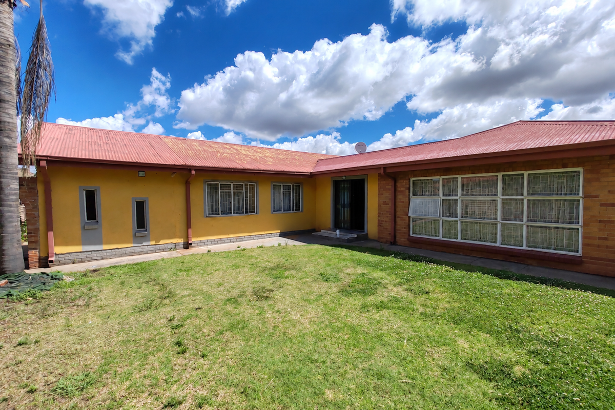 Rear view of a single-story house featuring a corrugated metal roof, brick and painted exterior walls, and large windows with security bars.