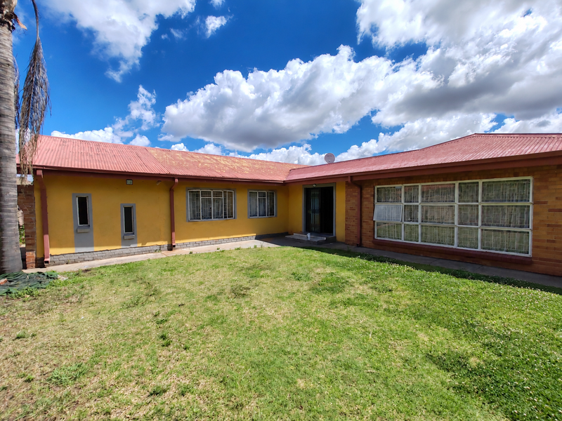 Rear view of a single-story house featuring a corrugated metal roof, brick and painted exterior walls, and large windows with security bars.