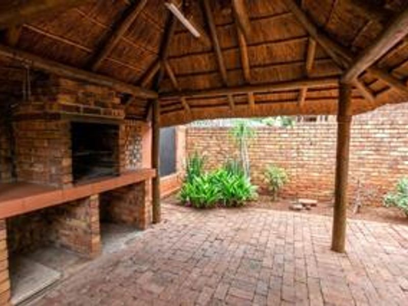 Covered outdoor patio area featuring a traditional thatched roof, exposed timber support beams, and a built-in brick braai, set against a private brick-walled garden.