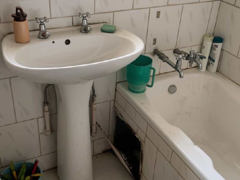 A bathroom featuring a pedestal basin and bathtub with tiled finishes, currently requiring maintenance and repair to the bath panel and tiling.