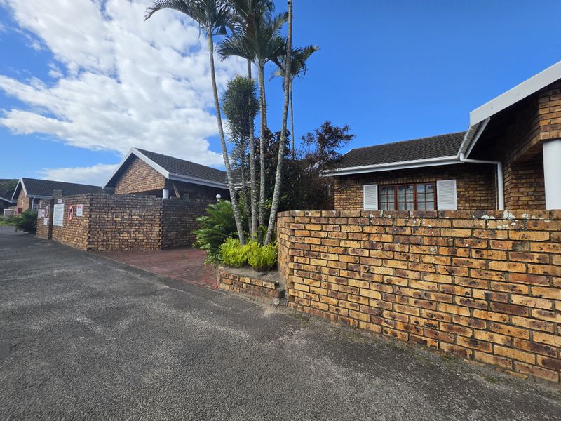Exterior view of a face brick townhouse complex featuring a paved driveway, boundary walls, and established palm trees under a clear blue sky.