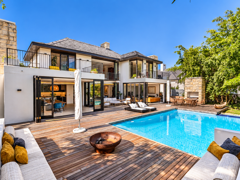 A luxurious outdoor entertainment area featuring a swimming pool, expansive timber decking, built-in seating, and a stone fireplace, seamlessly connected to the home's interior.