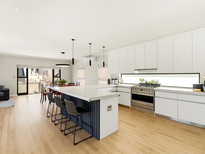 Modern open-plan kitchen and living area featuring a large island, stainless steel gas cooktop, sleek white cabinetry, and light wood-look flooring with seamless outdoor access.