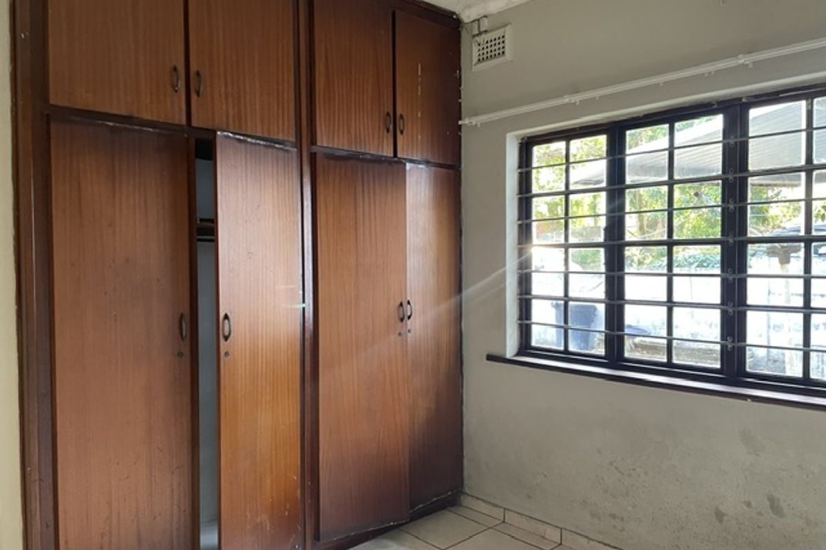 A bedroom featuring built-in wooden cupboards, tiled flooring, and a window with security bars, requiring cosmetic updates to the walls and cabinetry.