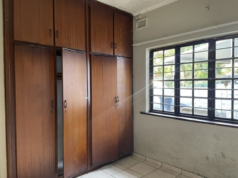 A bedroom featuring built-in wooden cupboards, tiled flooring, and a window with security bars, requiring cosmetic updates to the walls and cabinetry.