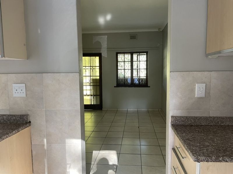 An open-plan living area viewed from the kitchen, featuring tiled flooring, neutral walls, and direct access to an outdoor space through a security gate.