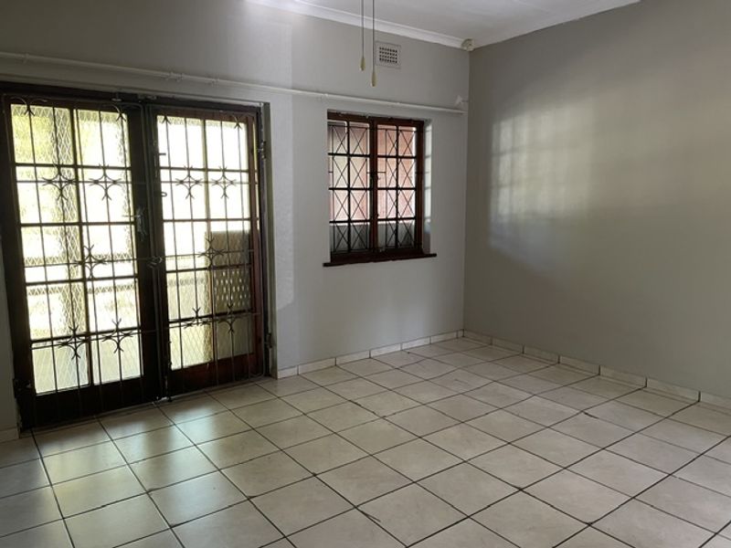 A bright, empty living room featuring tiled flooring, a ceiling fan, and secure double doors leading to an outdoor area.