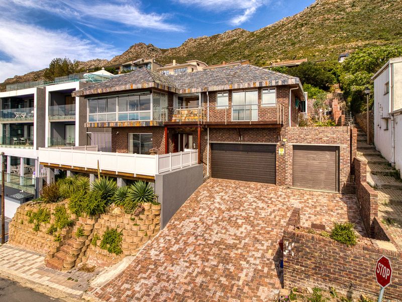 A multi-level brick house featuring a paved driveway, double garage, and an elevated balcony, set against a mountain backdrop.
