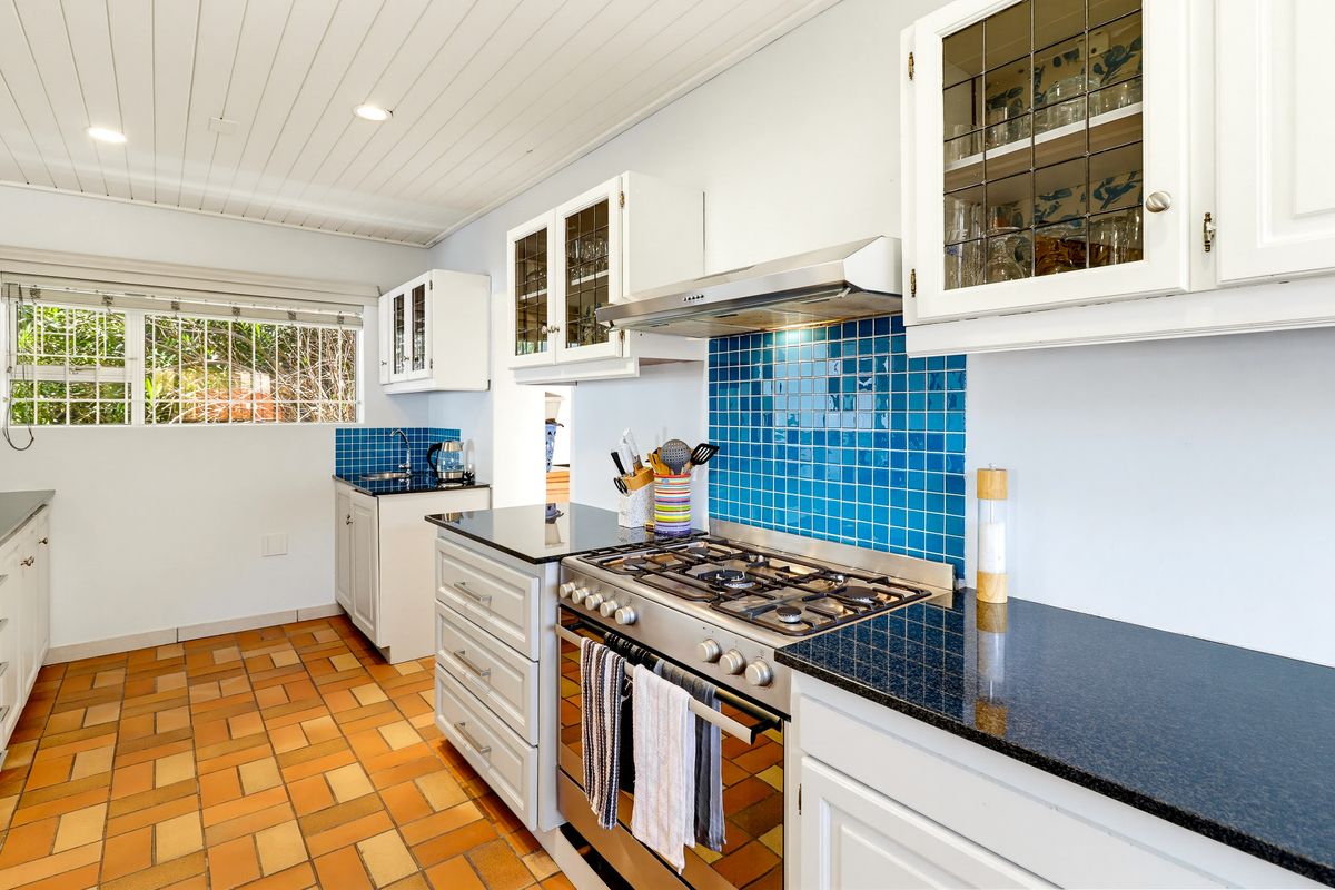 A well-maintained kitchen featuring granite countertops, a gas stove with extractor fan, blue tiled backsplash, and classic white cabinetry with terracotta flooring.