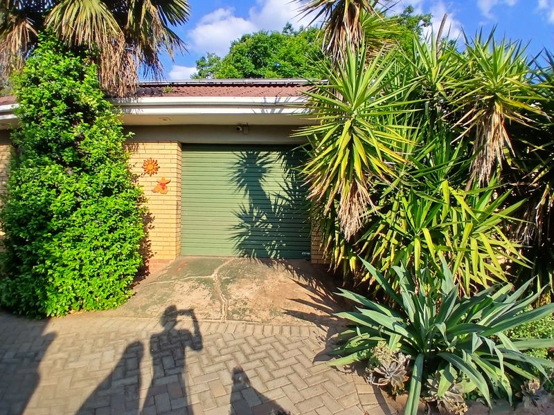 A single garage featuring a green roller shutter door, brick exterior, and a paved driveway surrounded by established garden landscaping.