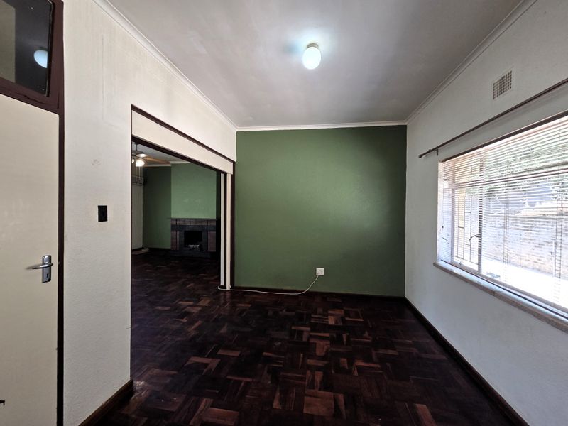 An empty living room featuring original parquet flooring, a green accent wall, and large windows with blinds, leading into an adjacent room with a fireplace.