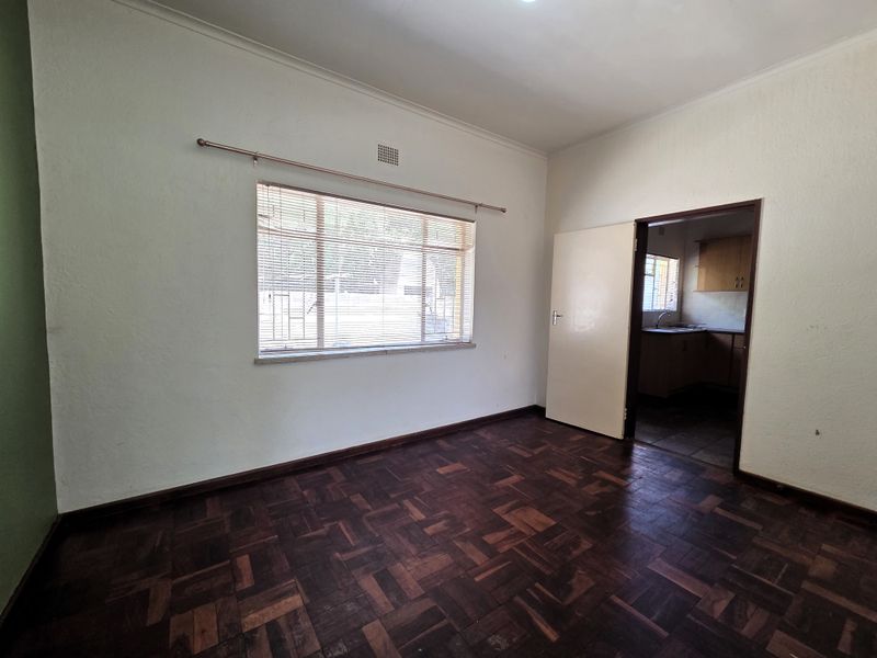An empty living room featuring original parquet flooring, coved cornicing, and a window with fitted venetian blinds, leading through to the kitchen area.