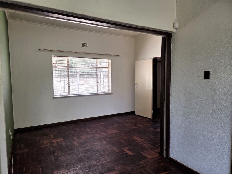 An empty living room featuring dark parquet flooring, a window with fitted blinds, and a security gate, offering a blank canvas for interior design.