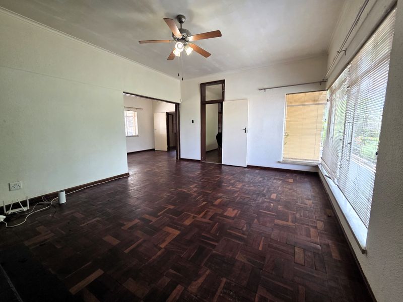 Spacious living area featuring original parquet flooring, a ceiling fan, and large windows with venetian blinds, offering a blank canvas for interior design.