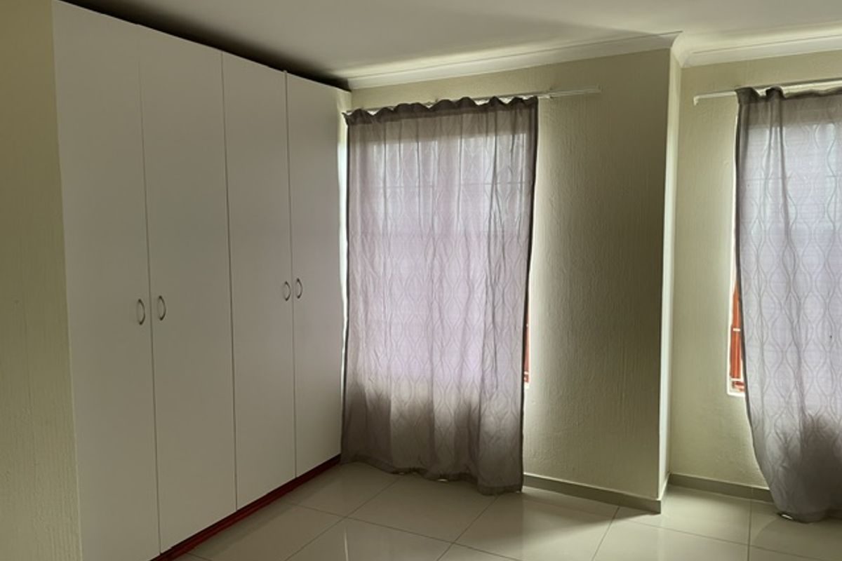 A bright, empty bedroom featuring neutral tiled flooring, white built-in cupboards, and large windows with curtains.
