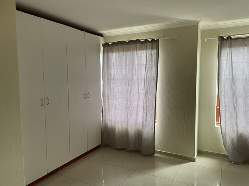 A bright, empty bedroom featuring neutral tiled flooring, white built-in cupboards, and large windows with curtains.