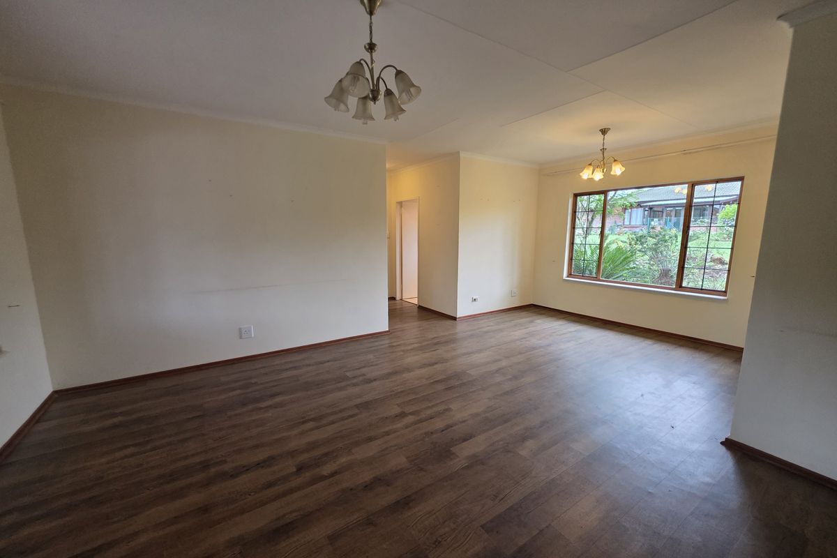 A spacious, empty living room featuring laminate flooring, coving, and large windows that provide natural light and a view of the garden.