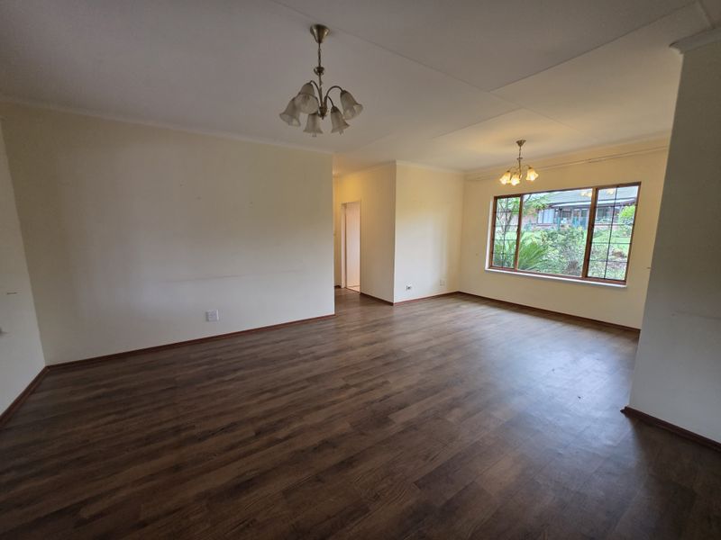 A spacious, empty living room featuring laminate flooring, coving, and large windows that provide natural light and a view of the garden.