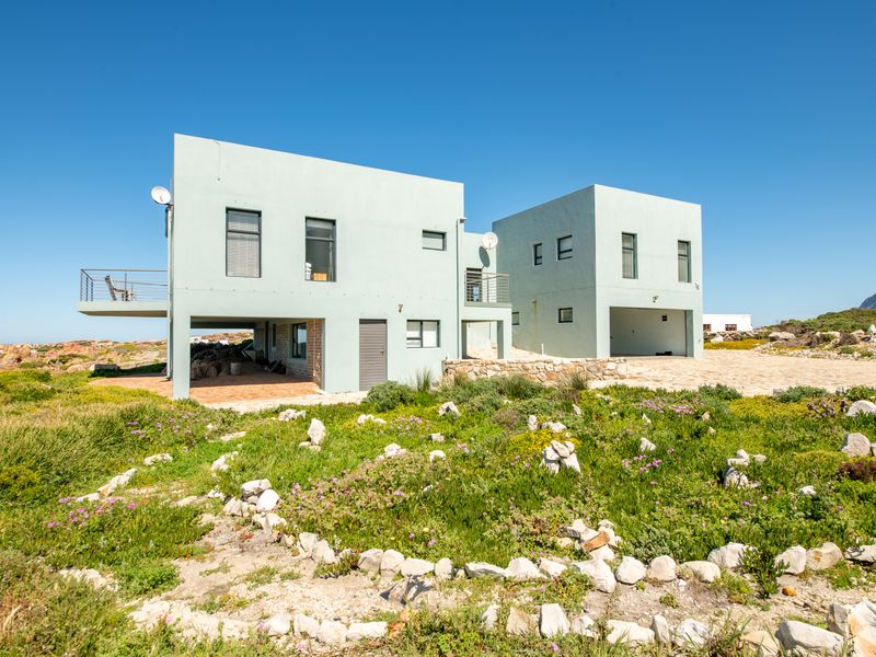 Modern two-story coastal residence featuring clean lines, a balcony, and a paved patio area set within a natural, landscaped garden environment.