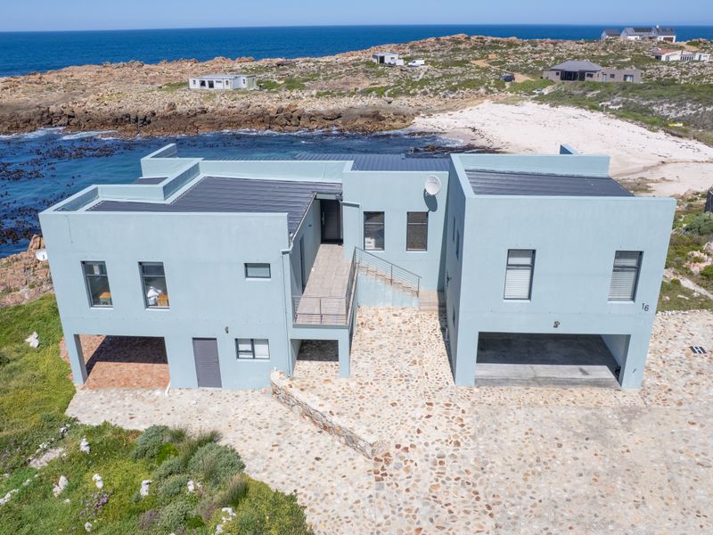 Aerial view of a modern coastal residence featuring a stone-paved driveway, integrated garage, and balcony, situated directly on the rocky shoreline.