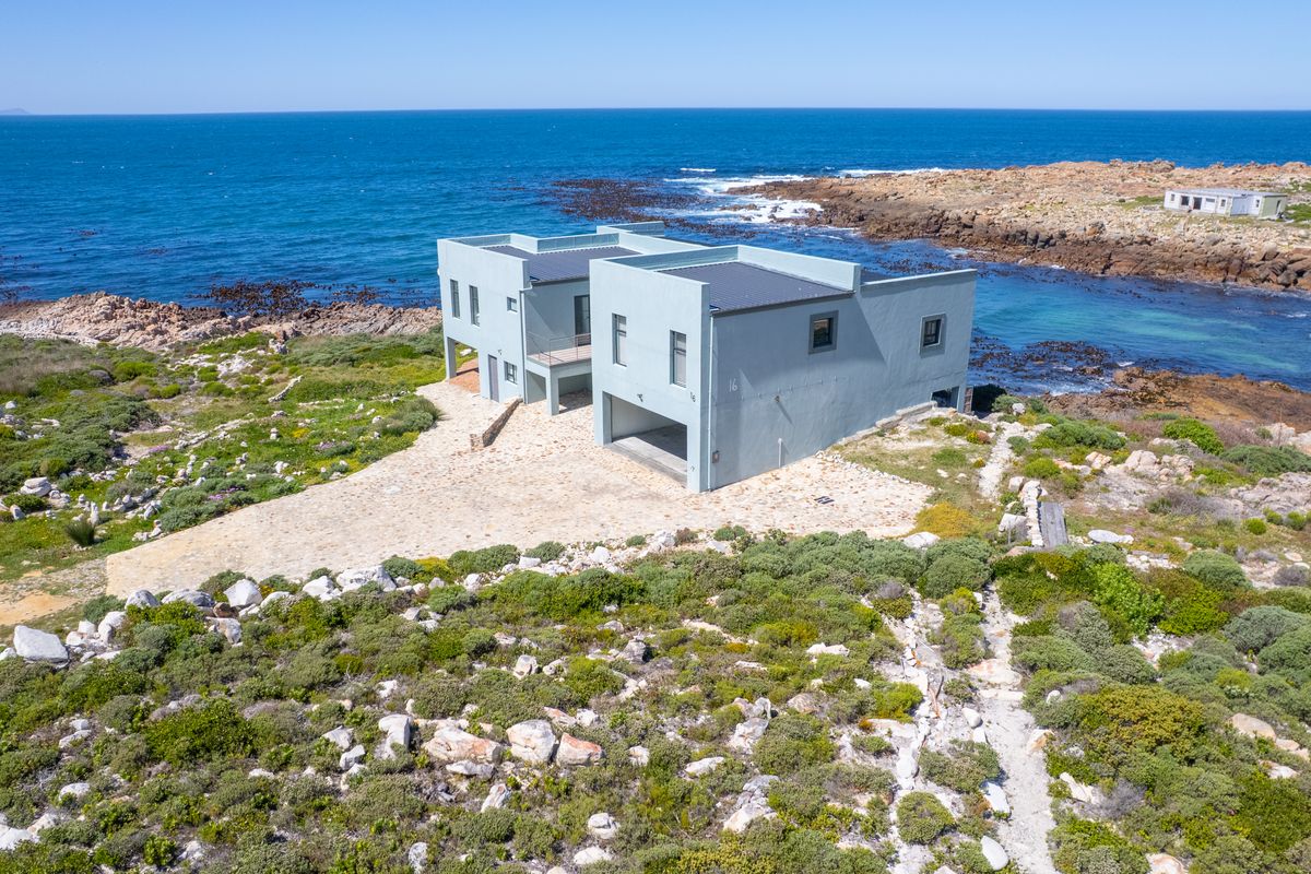 Aerial view of a modern coastal property featuring a flat roof design and stone paved driveway, situated directly on the rocky shoreline.
