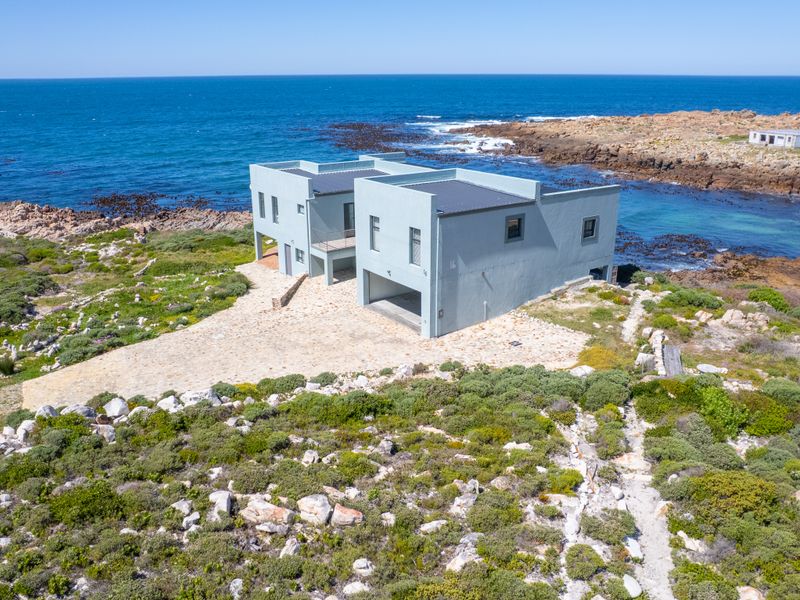 Aerial view of a modern coastal property featuring a flat roof design and stone paved driveway, situated directly on the rocky shoreline.