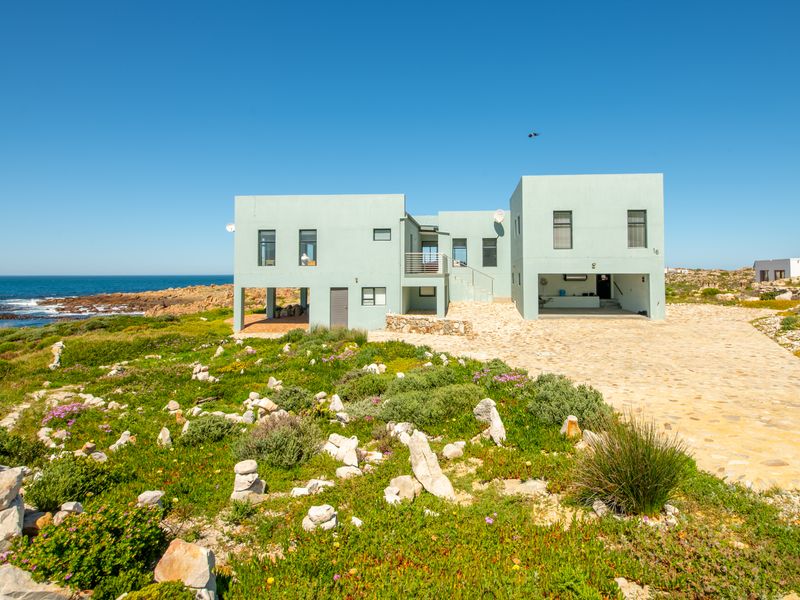 A modern coastal home featuring a stone-paved driveway, elevated balcony, and direct access to the rocky shoreline.