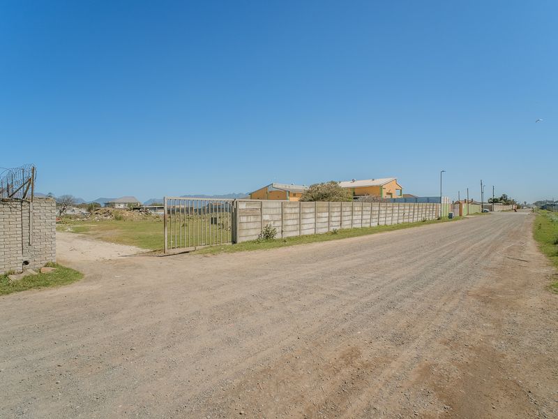Street view showing a gravel road access point to a vacant plot enclosed by precast concrete boundary walls and metal gates.