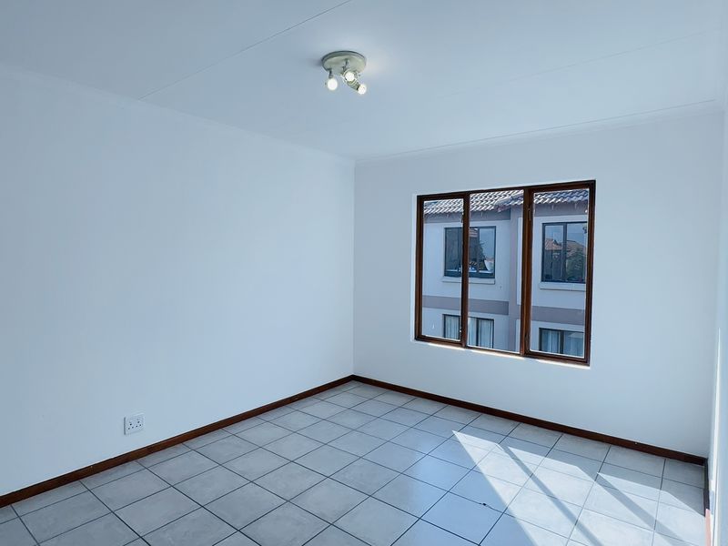 A bright, empty bedroom featuring neutral tiled flooring, white walls, and wooden-framed windows, providing a clean canvas for personalization.